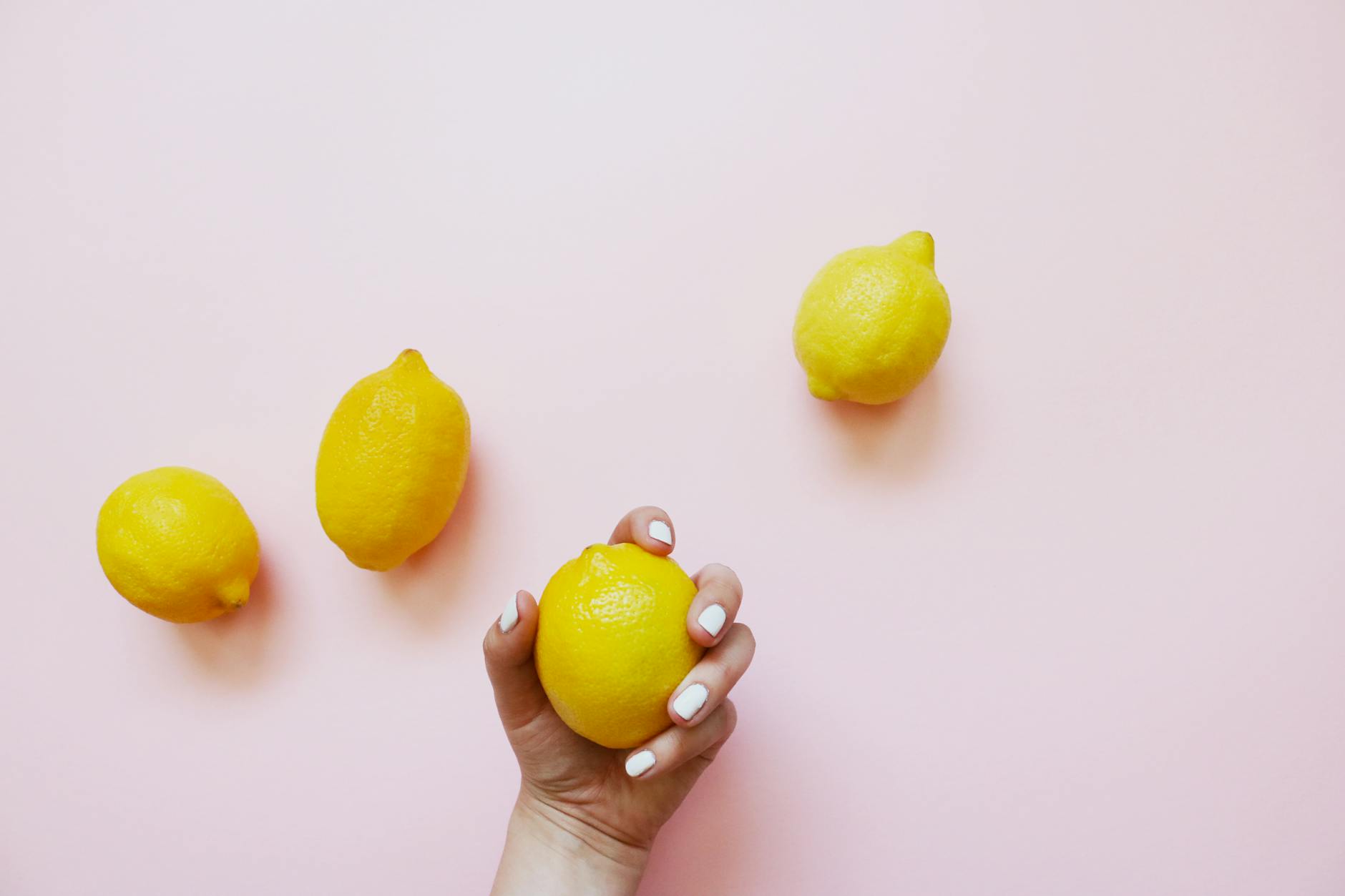 Une main tenant un citron sur un fond rose doux, entourée de trois citrons supplémentaires.