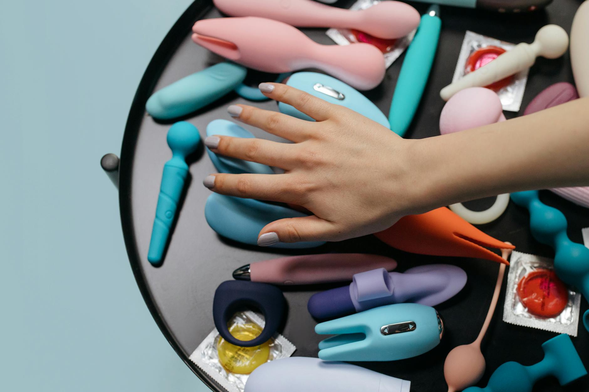 A hand reaching toward colorful vibrators arranged on a table, representing exploring pleasure without judgment.